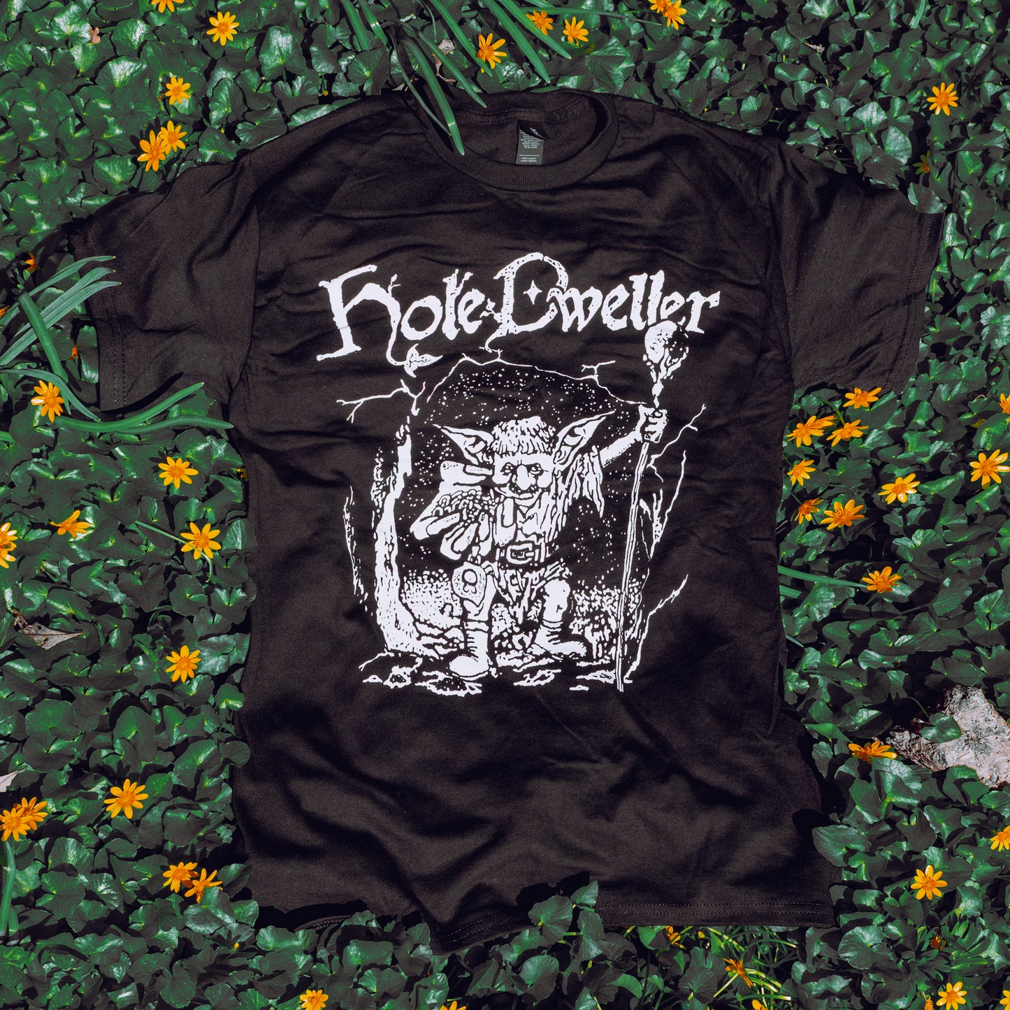 Black T-Shirt featuring an illustration of a goblin emerging from a cave, holding a staff with outreached hand. Arched above is the Hole Dweller logo. Printed with white ink. The shirt rests on a bed of green plants and flowers.