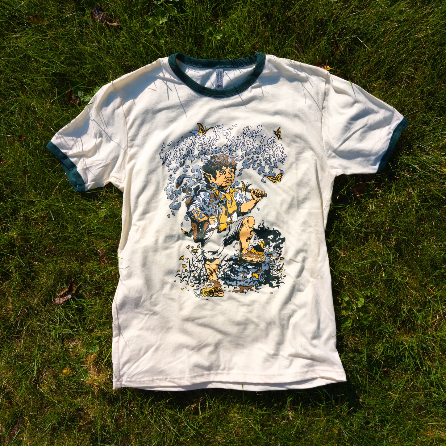 Natural cotton t-shirt with forest green ringers displaying a graphic of a hobbit holding a golf club with the title "Hole Dweller" above him which has formed from the smoke of the pipe the hobbit is holding. The shirt is resting on the grass.