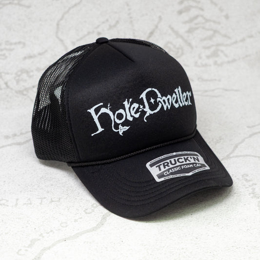 Black trucker cap with white 'Hole Dwellers' logo on a textured white background