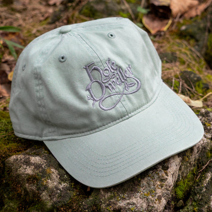 A light sage green baseball cap with a grey embroidered script logo on the front, resting on a mossy rock outdoors.