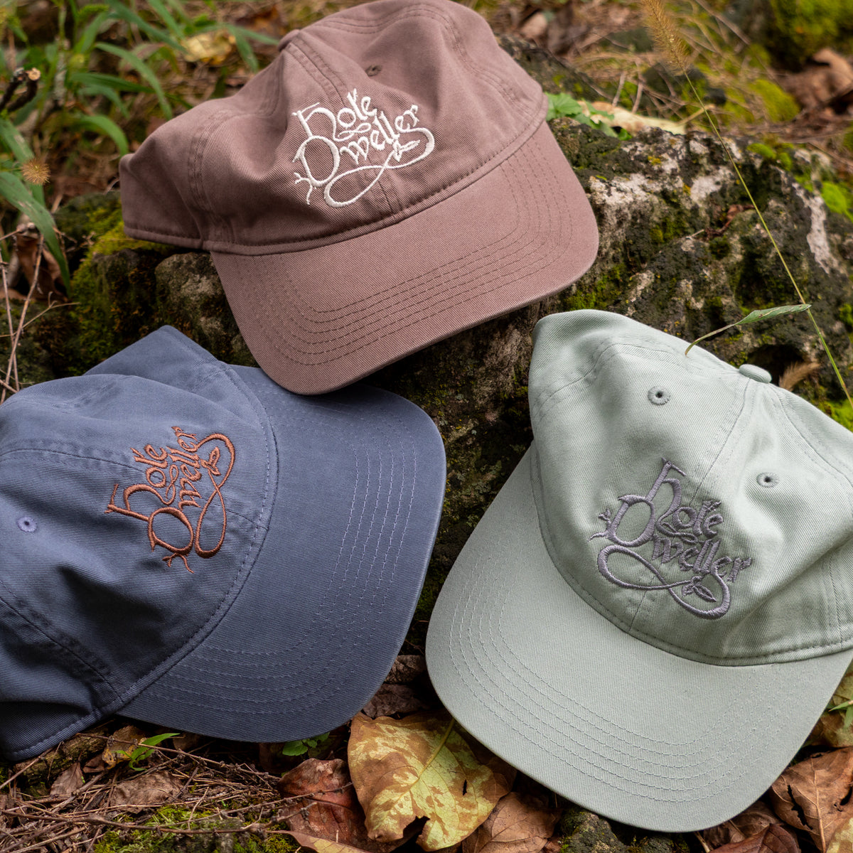 Three baseball caps in brown, navy, and sage green are arranged on a mossy log outdoors. Each cap features a curved brim and an embroidered script logo on the front panel.