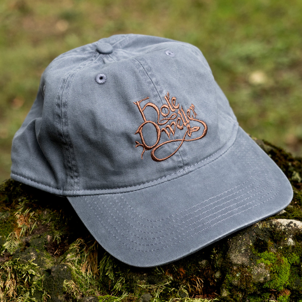 A navy blue cotton baseball cap with a brown embroidered script logo on the front, resting on a mossy log outdoors with a blurred green forest background.