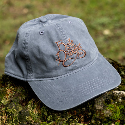 A navy blue cotton baseball cap with a brown embroidered script logo on the front, resting on a mossy log outdoors with a blurred green forest background.