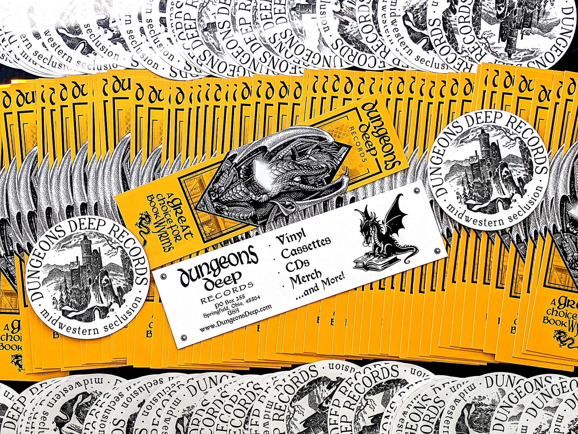 Yellow Dungeons Deep Records Bookmarks, and white round stickers fanned out across the image. The bookmarks show an image of a dragon reading a book, with the reverse side stating "Dungeons Deep Records - Vinyl, Cassettes, CDs, Merch and More!"