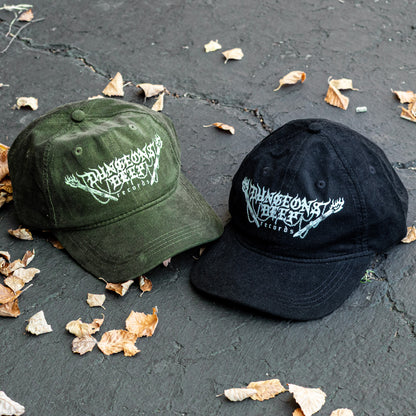 Two corduroy baseball caps, one green and one black, with a Dungeons Deep Records logo on a dark surface with scattered leaves.