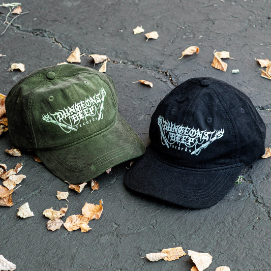 Two corduroy baseball caps, one green and one black, with a Dungeons Deep Records logo on a dark surface with scattered leaves.