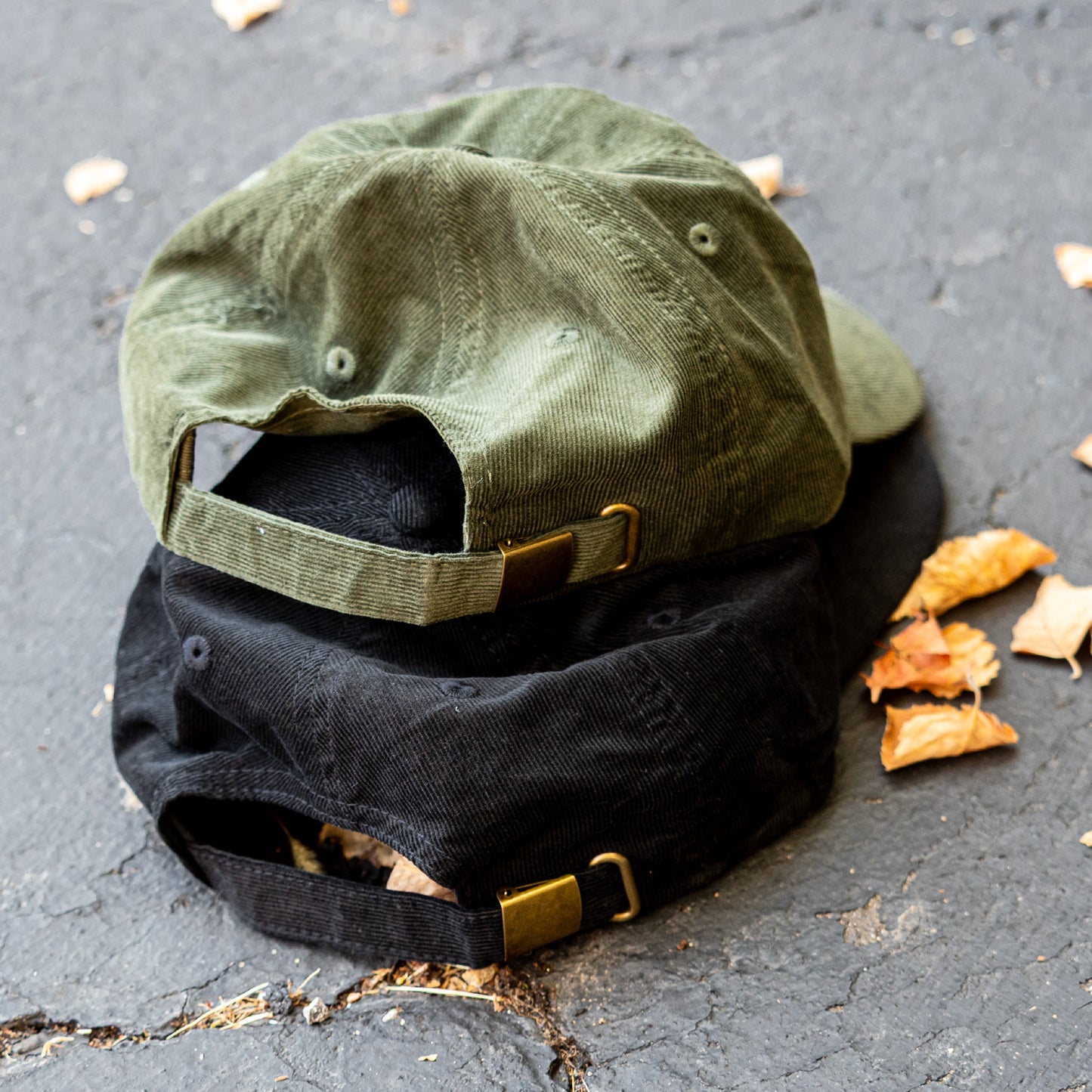 The back of two baseball caps, one green and one black, on a textured surface with dried leaves.