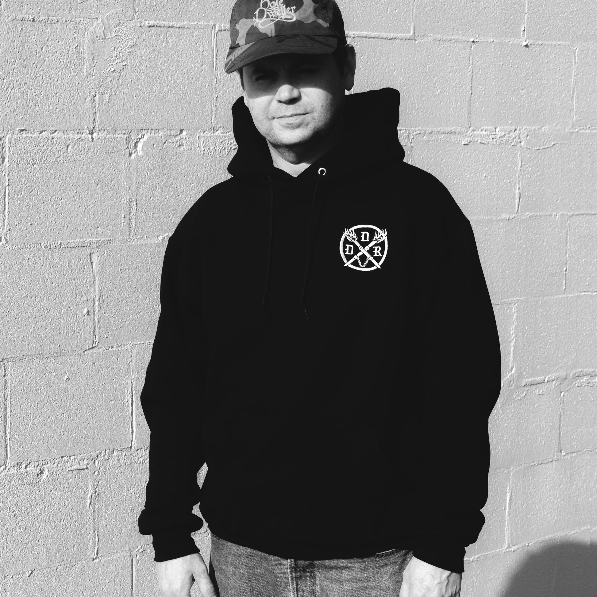 Person wearing a black hoodie with a white embroidered Dungeons Deep Records emblem against a gray brick wall