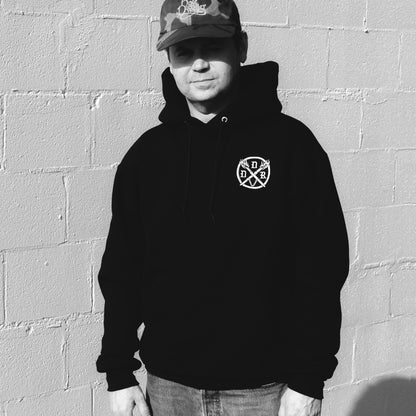 Person wearing a black hoodie with a white embroidered Dungeons Deep Records emblem against a gray brick wall