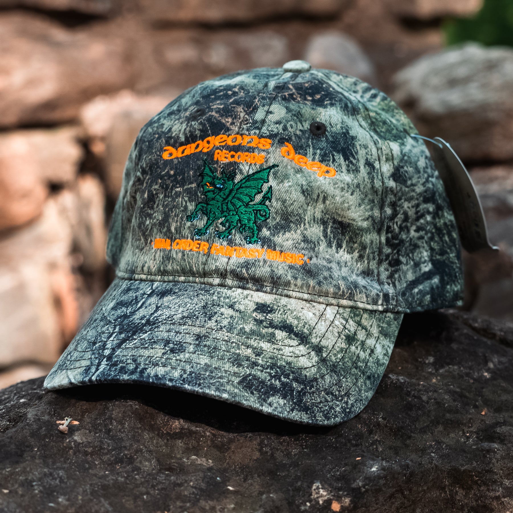 A Mossy Oak camouflage baseball cap on a stone surface. It is embroidered with a green dragon and the phrase; "Dungeons Deep Records - Mail Order Fantasy Music" in Neon Orange stitching.