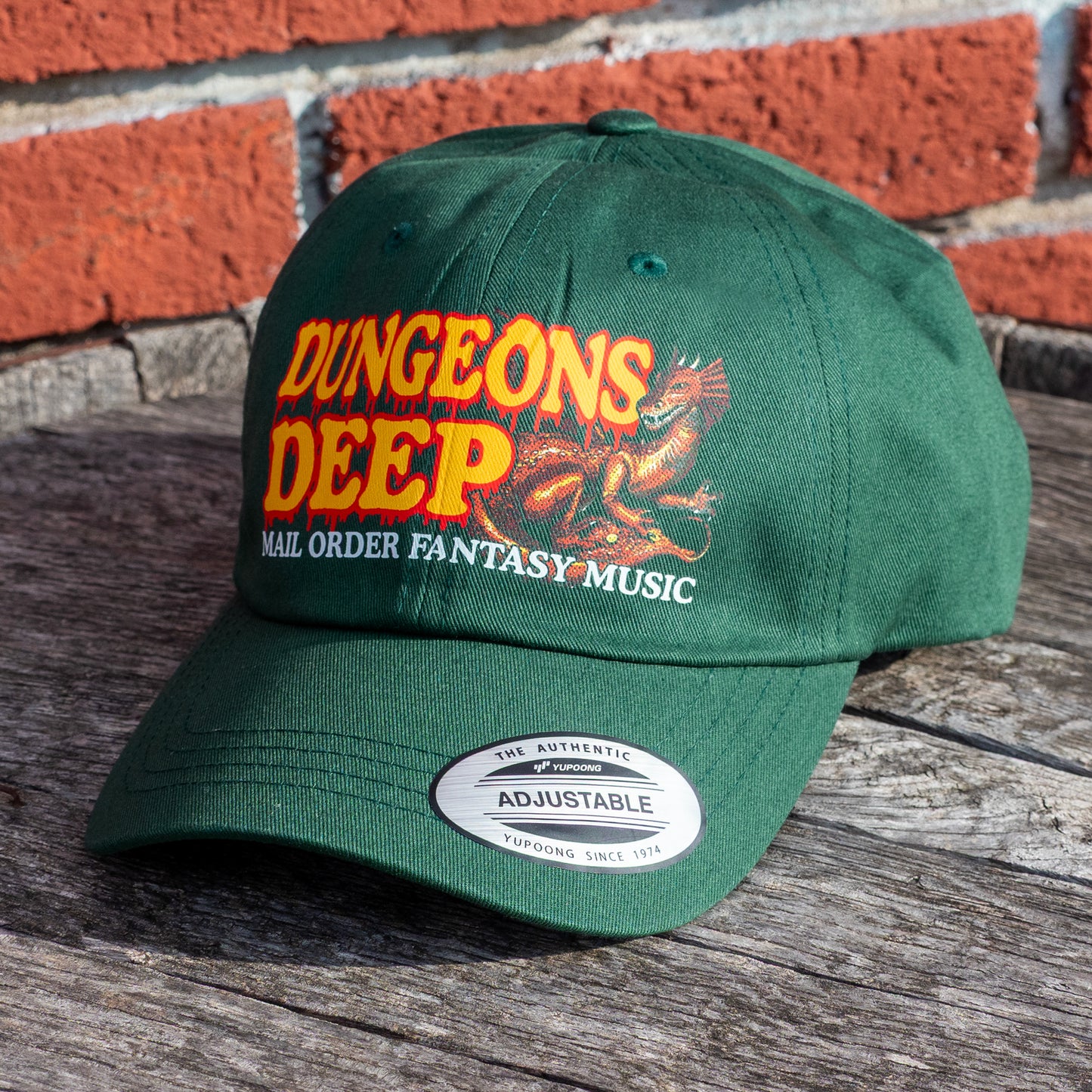 A green dad hat embellished with the Dungeons Deep Records "Mail Order Fantasy Music" Dragon emblem on a wooden backdrop.