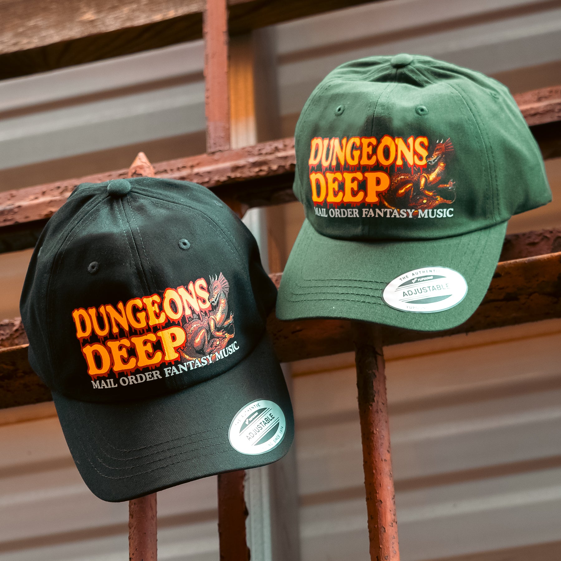 A black and green dad hat embellished with the Dungeons Deep Records "Mail Order Fantasy Music" Dragon emblem hanging from iron bars.