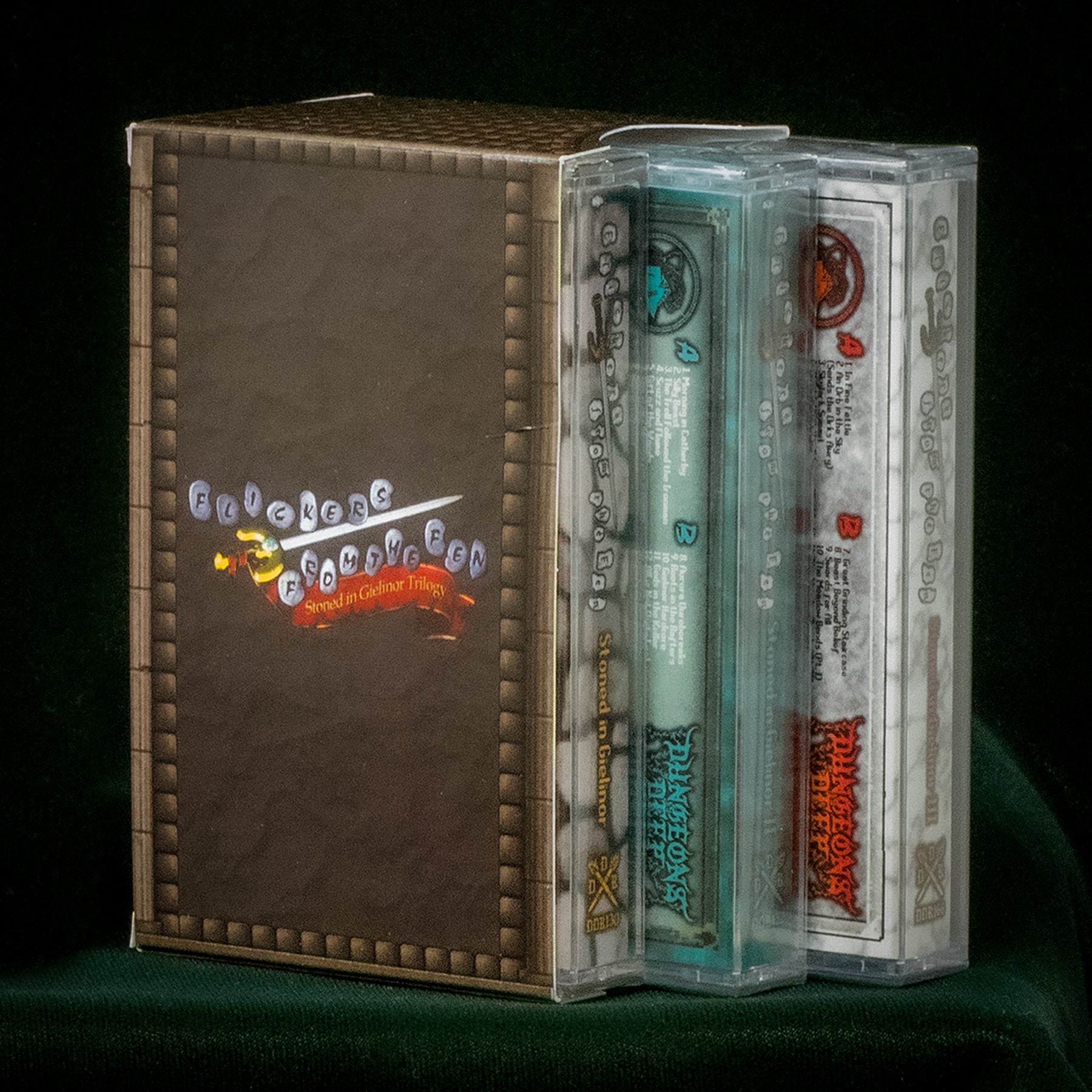 Three cassette tapes sit housed in a custom slipcase. The slipcase is brown and features the Flickers From the Fen logo in a Runsecape style font. The cassettes are from the Flickers From the Fen Stoned in Gielinor Trilogy.