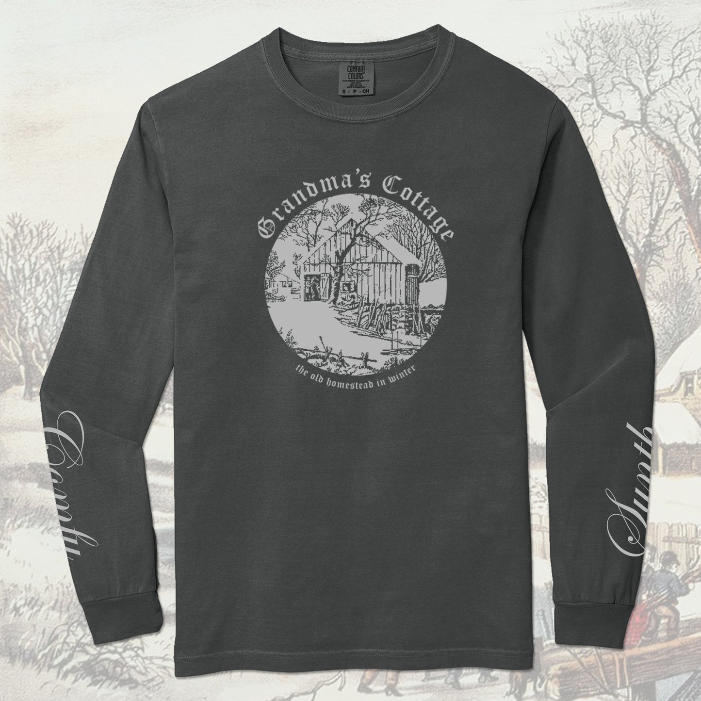 Dark gray long-sleeve shirt with a winter cabin scene graphic design. The left sleeve reads Synth, the right sleeve reads Comfy - both in cursive script. On a winter-themed background.