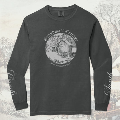 Dark gray long-sleeve shirt with a winter cabin scene graphic design. The left sleeve reads Synth, the right sleeve reads Comfy - both in cursive script. On a winter-themed background.