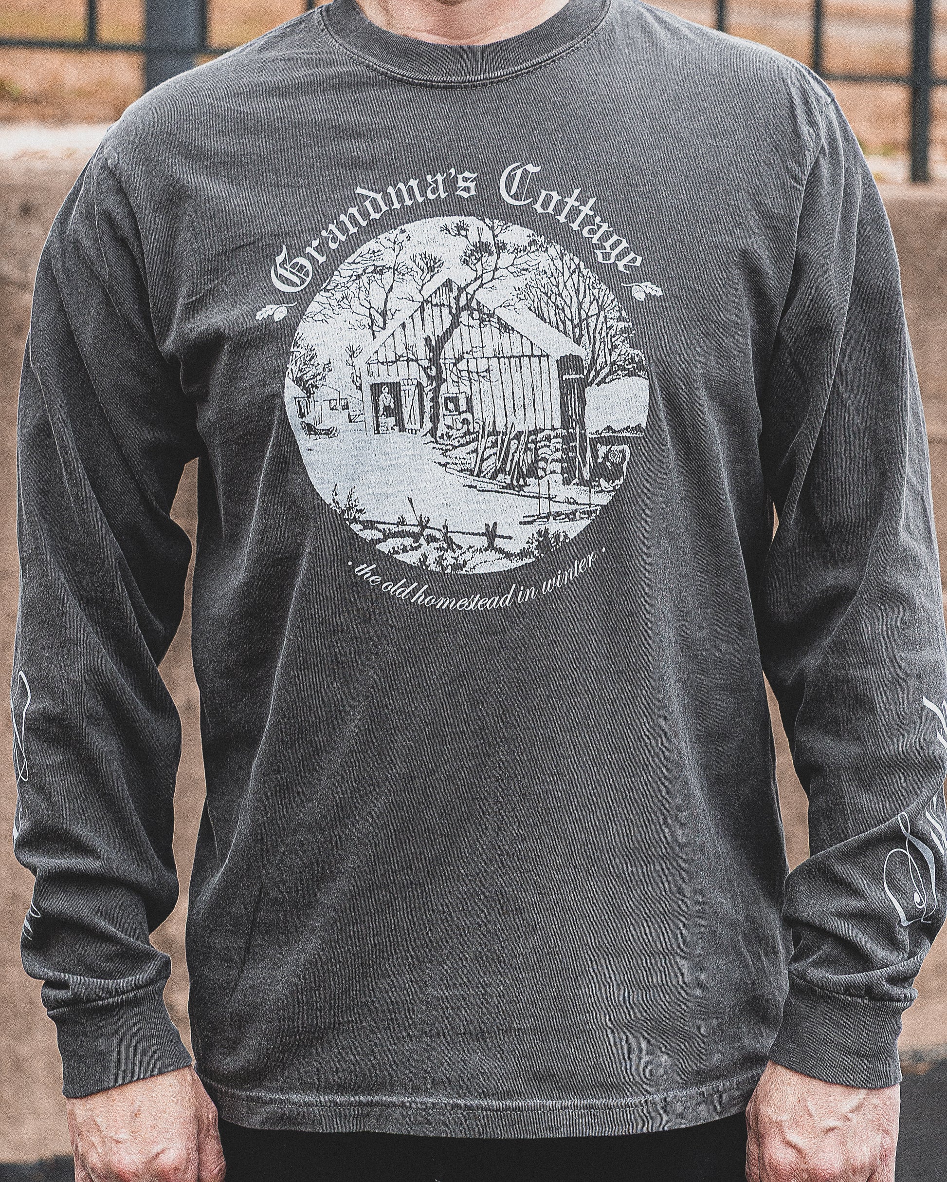 A man wears a grey comfort colors long sleeve t-shirt. The front of the shirt has the Grandma's Cottage logo with an illustration of an old cabin.