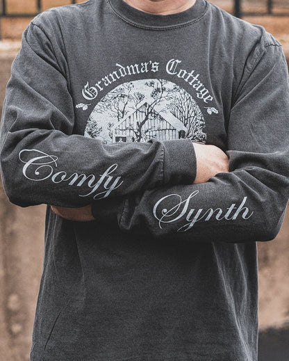 A man wears a grey comfort colors long sleeve t-shirt. His arms are crossed displaying the screen printed phrase Comfy Synth spanning both sleeves. The front of the shirt has the Grandma's Cottage logo with an illustration of an old cabin.