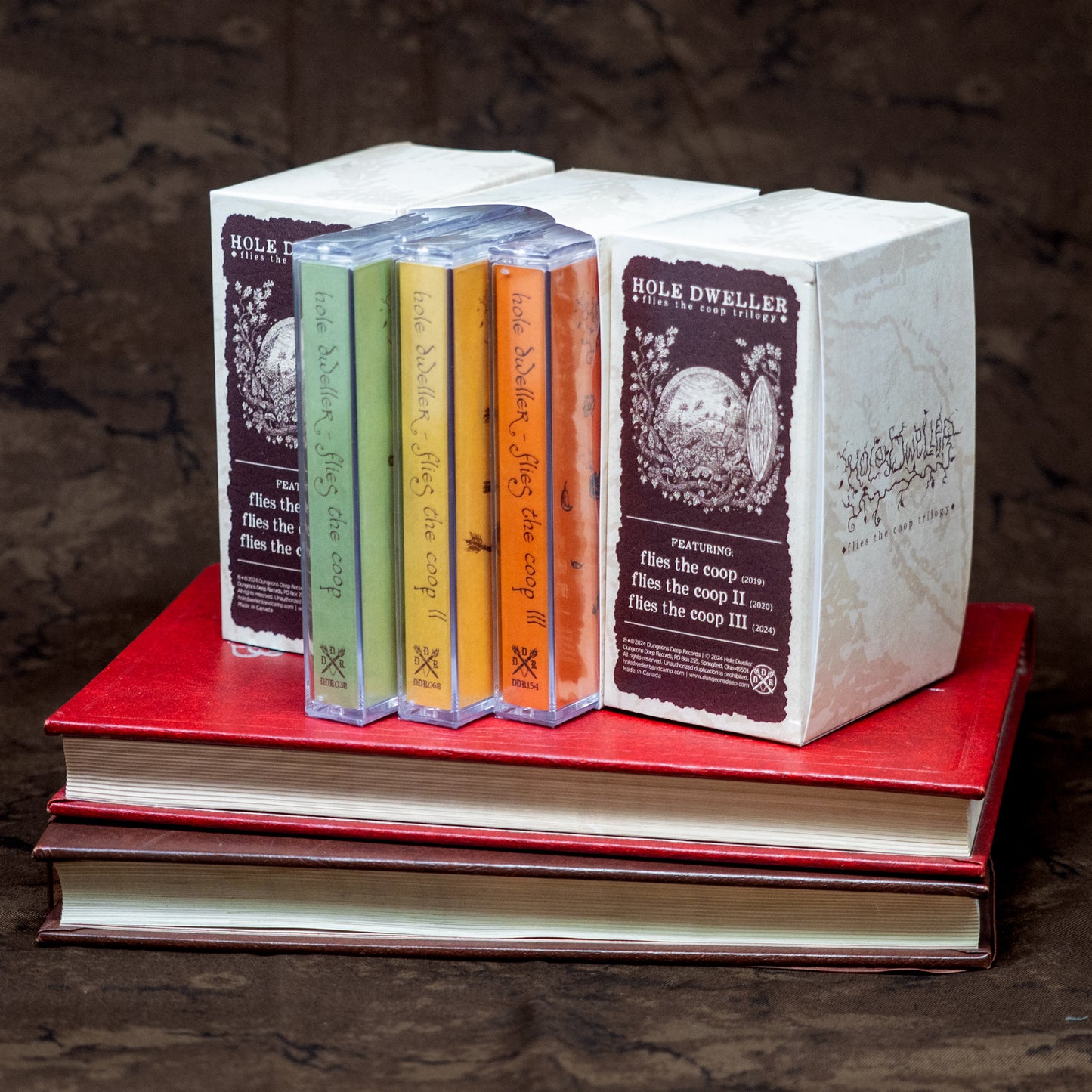 Three Hole Dweller "flies the coop" cassette trilogy boxsets sit next to one another, displaying all sides of the slipcase. The slipcase reads "Hole Dweller Flies the Coop Trilogy, Featuring Flies the Coop I, III and III. The slipcases rest on two old books.