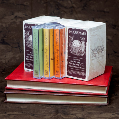 Three Hole Dweller "flies the coop" cassette trilogy boxsets sit next to one another, displaying all sides of the slipcase. The slipcase reads "Hole Dweller Flies the Coop Trilogy, Featuring Flies the Coop I, III and III. The slipcases rest on two old books.