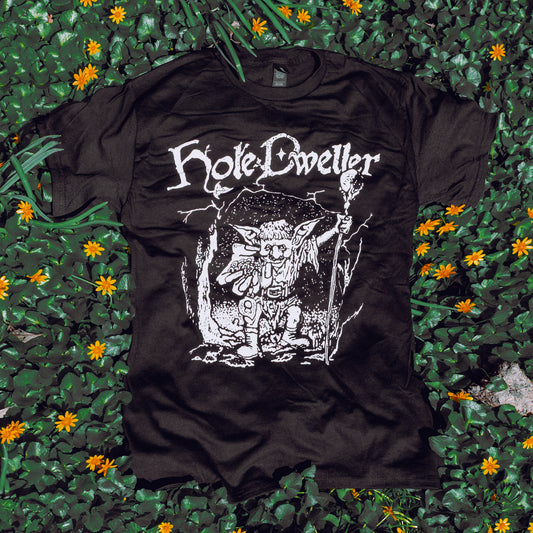 Black T-Shirt featuring an illustration of a goblin emerging from a cave, holding a staff with outreached hand. Arched above is the Hole Dweller logo. Printed with white ink. The shirt rests on a bed of green plants and flowers.