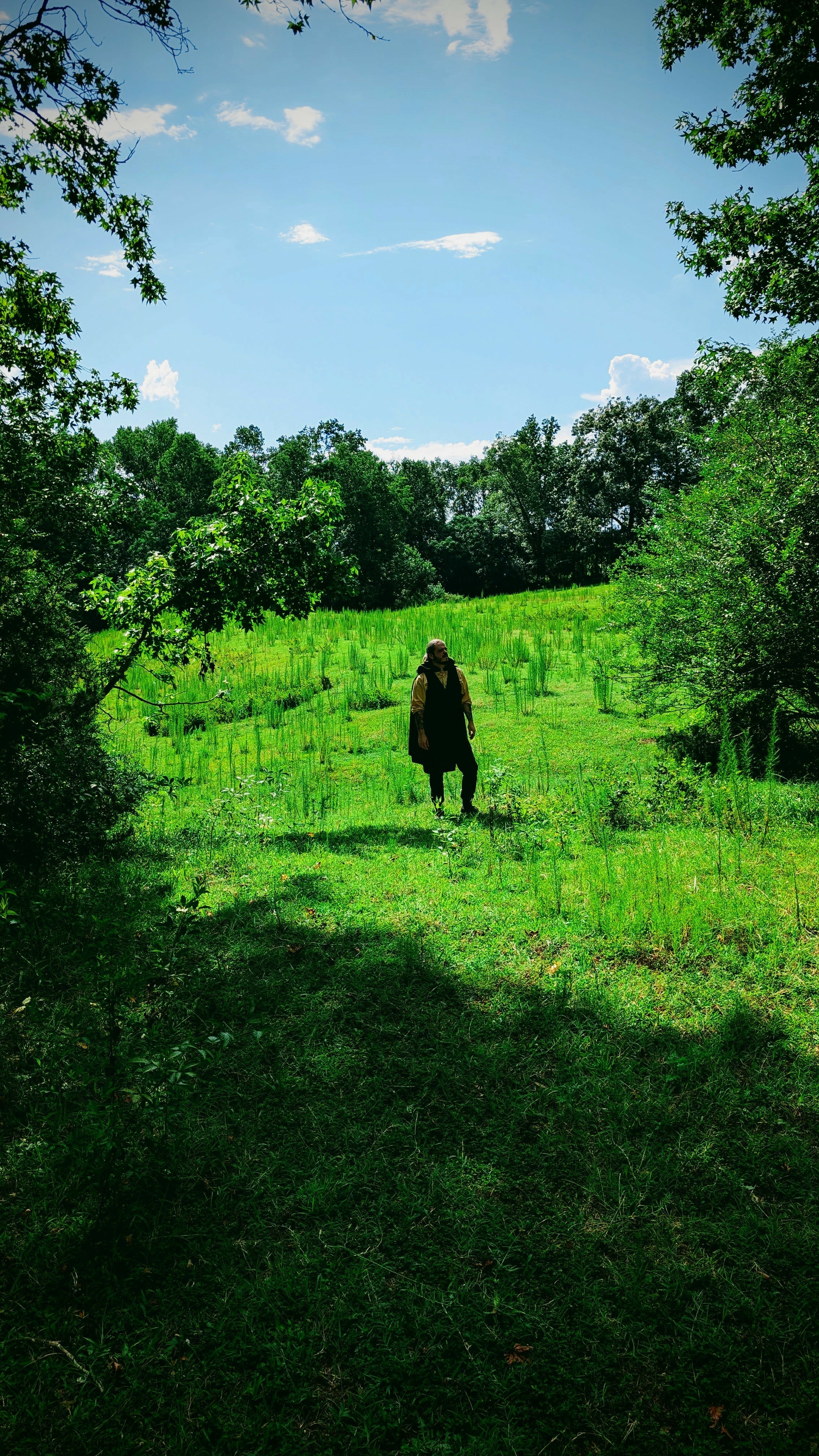Tim Rowland of Hole Dweller wears a green cape. He is walking through a green field on a sunny day.