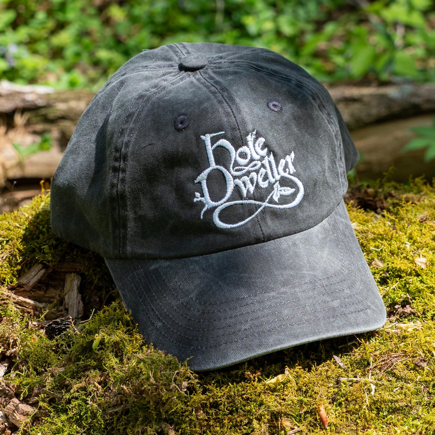 Baseball cap with embroidered  'Hole Dweller' logo on a mossy surface. The cap is a distressed grey color with silver stitching.