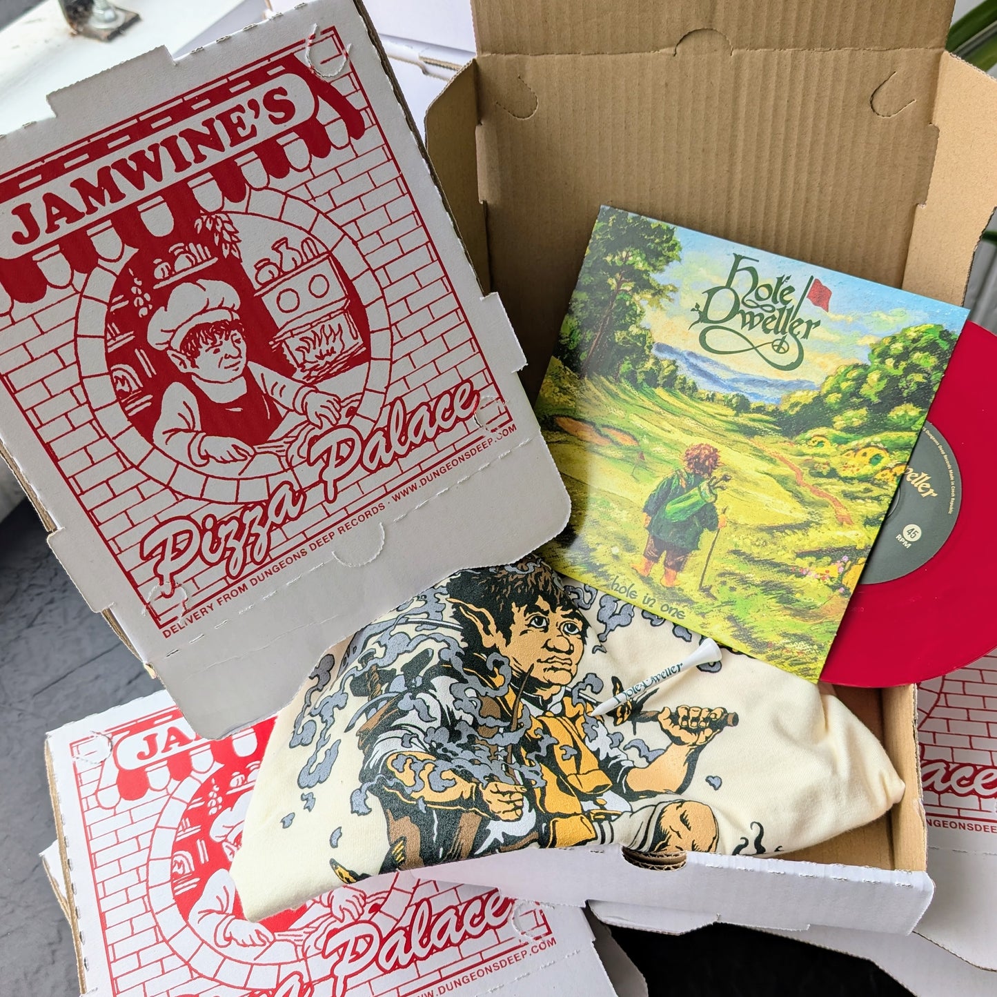 Open pizza box with 'Jamwine's Pizza Palace' branding holding a Hole Dweller T-Shirt, 7" record and golf tee.