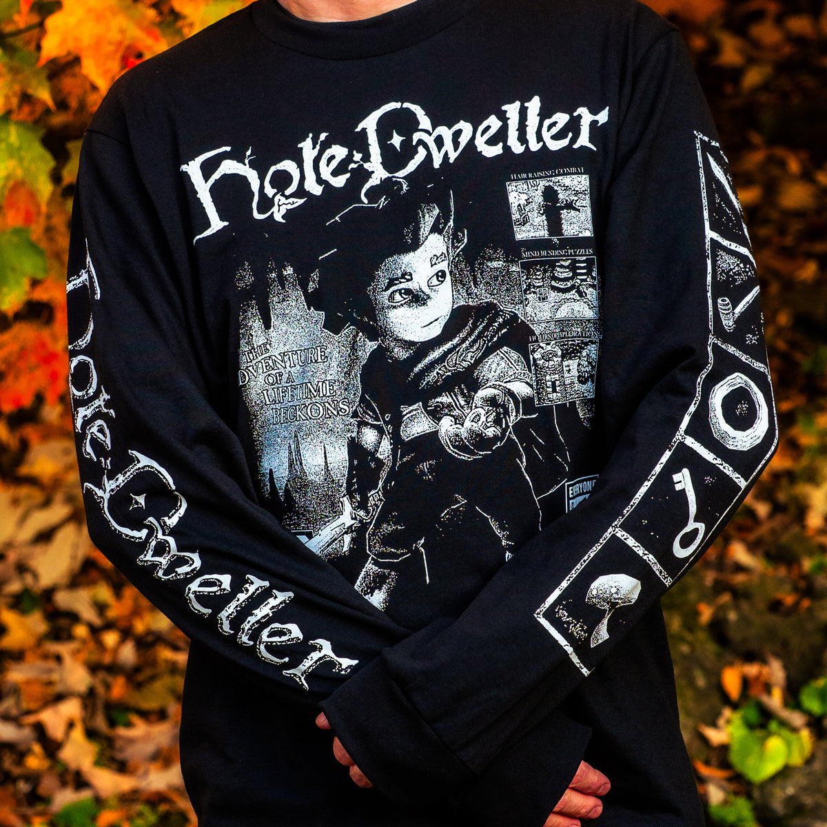 A man wearing a black long sleeve tshirt with white print. The front has an image of a retro PS2 style hobbit holding a ring, and the sleeves have images of video game inventory items, and the hole dweller logo.