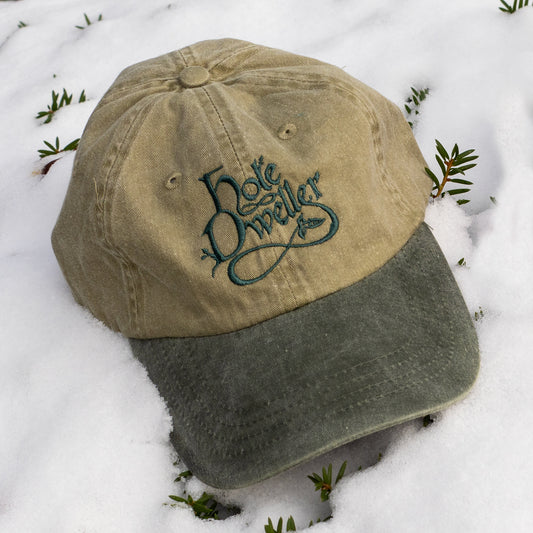A unisex baseball cap with a khaki crown and distressed forest green bill sits on a dusting of snow. The Hole Dweller logo is embroidered on the crown in forest green stitching.