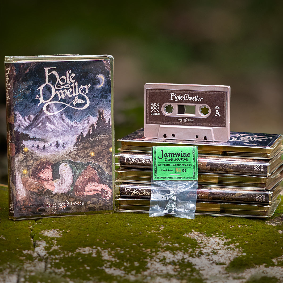 A themed setup featuring a 'Hole Dweller' cassette tape, a matching cassette cover, several stacked cassette cases, and a small clear bag of Jamwine  pewter miniature placed on a mossy surface with a blurred outdoor background.
