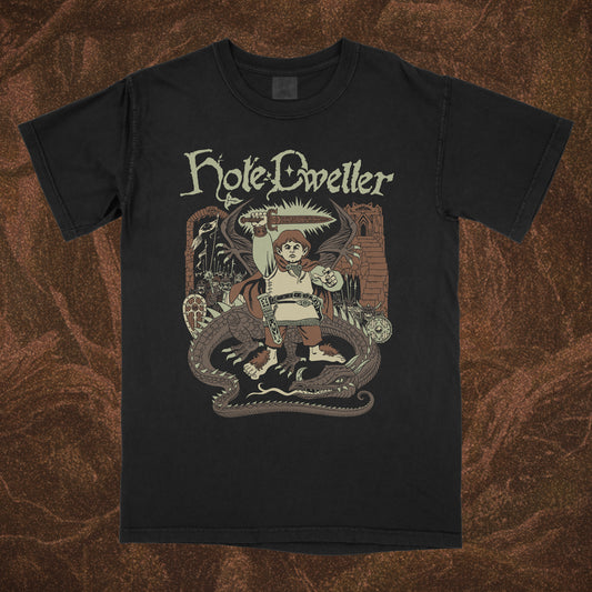 Black short-sleeve Comfort Colors shirt with 'Hole Dweller' graphic design featuring a hobbit holding a sword to the sky. The right sleeve reads Hole Dweller vertically, the left sleeve features a sword. The image is printed in tan, green and brown. The shirt is against a brown textured backdrop. 