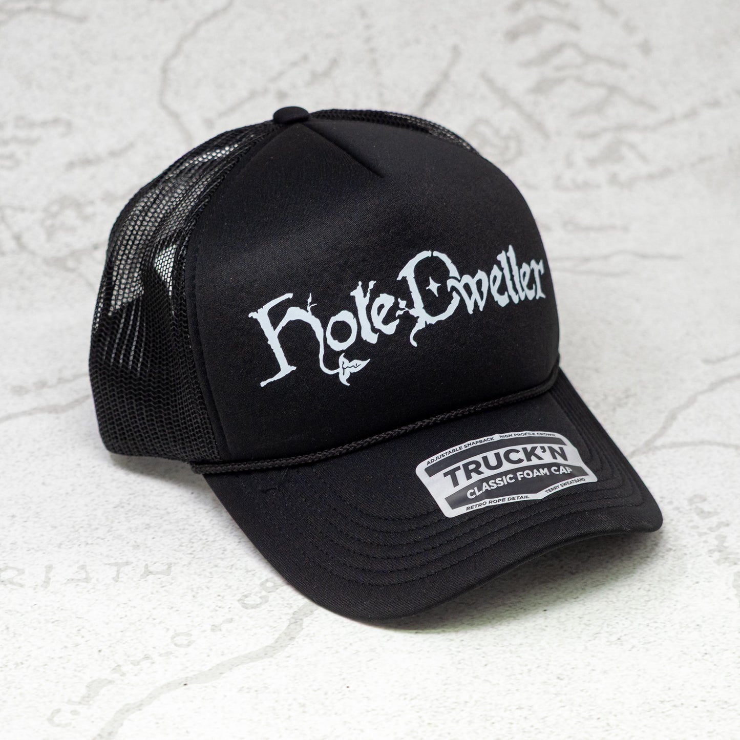 Black trucker cap with white 'Hole Dwellers' logo on a textured white background