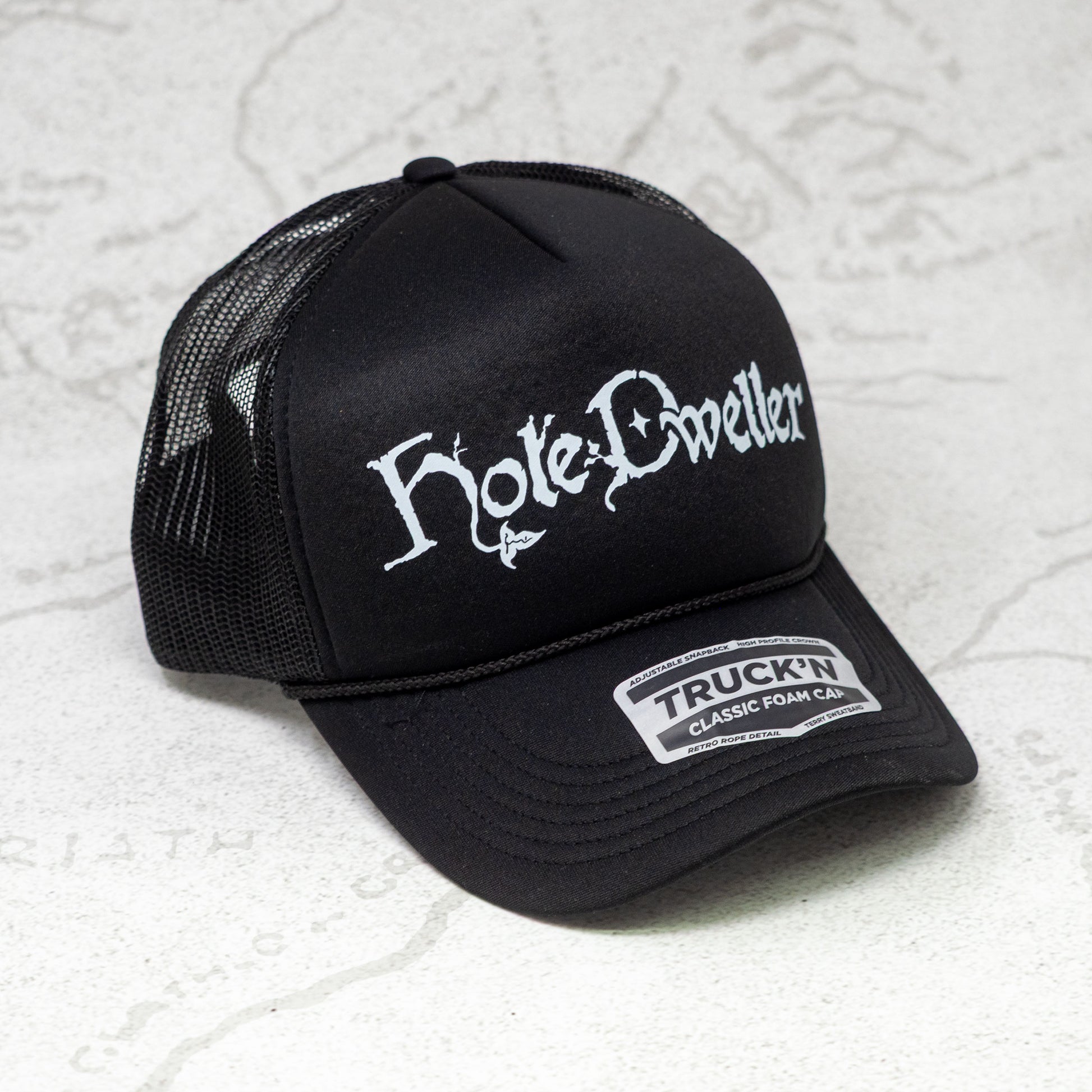 Black trucker cap with white 'Hole Dwellers' logo on a textured white background