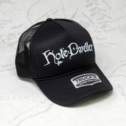 Black trucker cap with white 'Hole Dwellers' logo on a textured white background