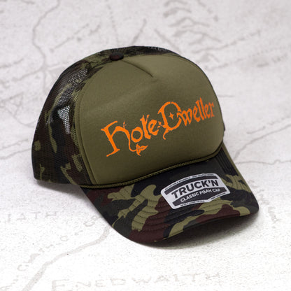 Camouflage cap with orange 'Hole Dweller' text on a textured white background