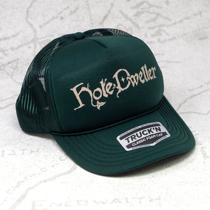 Forest green trucker cap with tan 'Hole Dweller' text on a textured white background