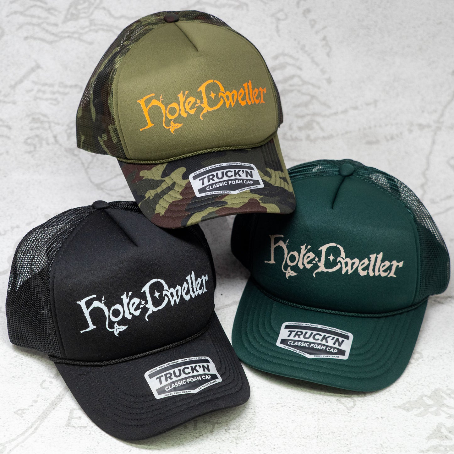 Three 'Hole-Dweller' branded caps on a textured white background. One hat is Camo with an Orange Hole Dweller logo, one is Black with a white logo, and the third is forest green with a tan logo.