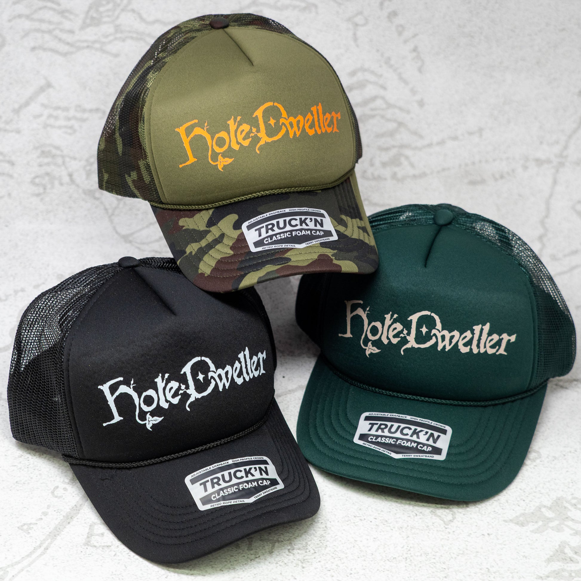 Three 'Hole-Dweller' branded caps on a textured white background. One hat is Camo with an Orange Hole Dweller logo, one is Black with a white logo, and the third is forest green with a tan logo.