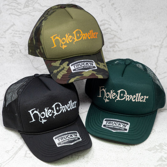 Three 'Hole-Dweller' branded caps on a textured white background. One hat is Camo with an Orange Hole Dweller logo, one is Black with a white logo, and the third is forest green with a tan logo.