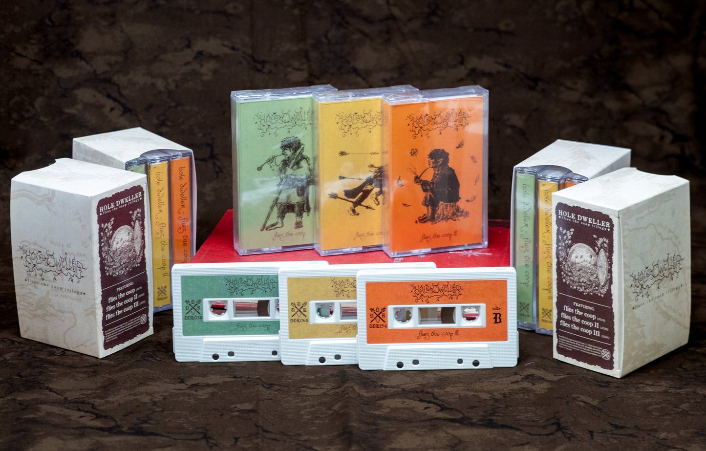 Three Hole Dweller "flies the coop" cassette trilogy boxsets sit next to one another, displaying all sides of the slipcase and the front of the cassette shells. The slipcase reads "Hole Dweller Flies the Coop Trilogy, Featuring Flies the Coop I, III and III. The slipcases rest on two old books on a brown background.