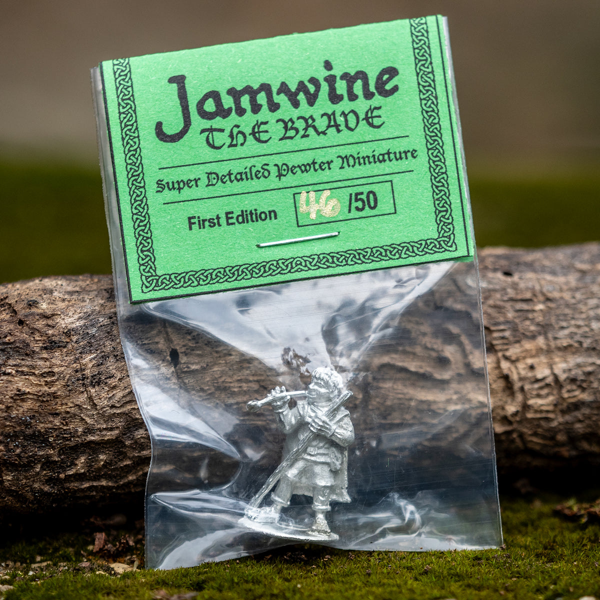 A packaged miniature figurine labeled 'Jamwine The Brave' in a clear plastic bag with a green paper header card, resting on moss beside a log outdoors.