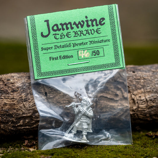A packaged miniature figurine labeled 'Jamwine The Brave' in a clear plastic bag with a green paper header card, resting on moss beside a log outdoors.