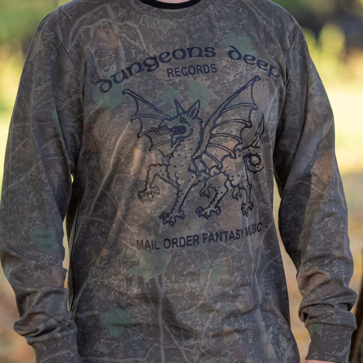 A man wearing a leafy Camouflaged long sleeve tshirt. In black print is a dragon, with the words Dungeons Deep Records arched across the top, and Mail order fantasy music below, on a blurry background.