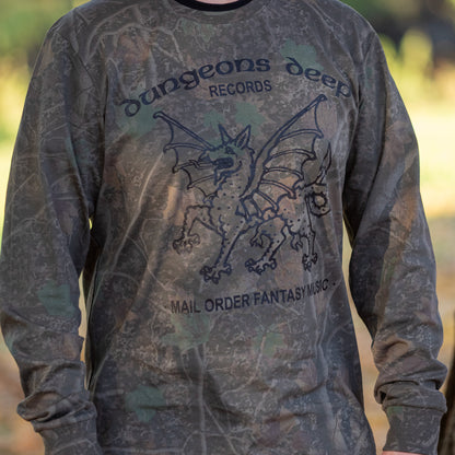 A man wearing a leafy Camouflaged long sleeve tshirt. In black print is a dragon, with the words Dungeons Deep Records arched across the top, and Mail order fantasy music below, on a blurry background.