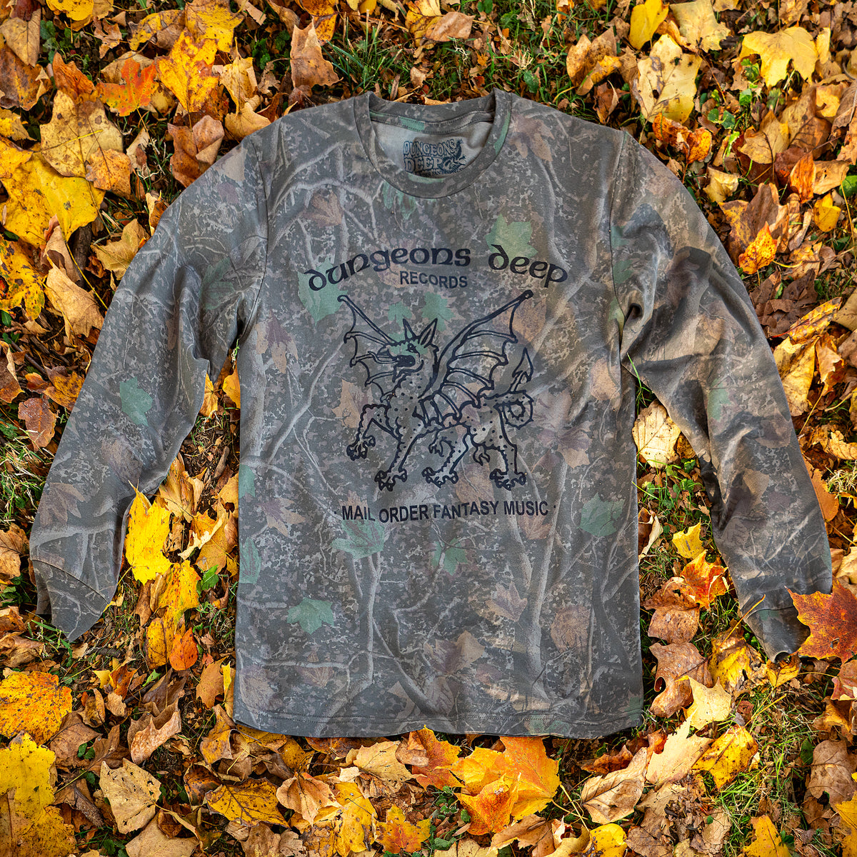 A leafy Camouflaged long sleeve tshirt laying on a pile of fall leaves. In black print is a dragon, with the words Dungeons Deep Records arched across the top, and Mail order fantasy music below.