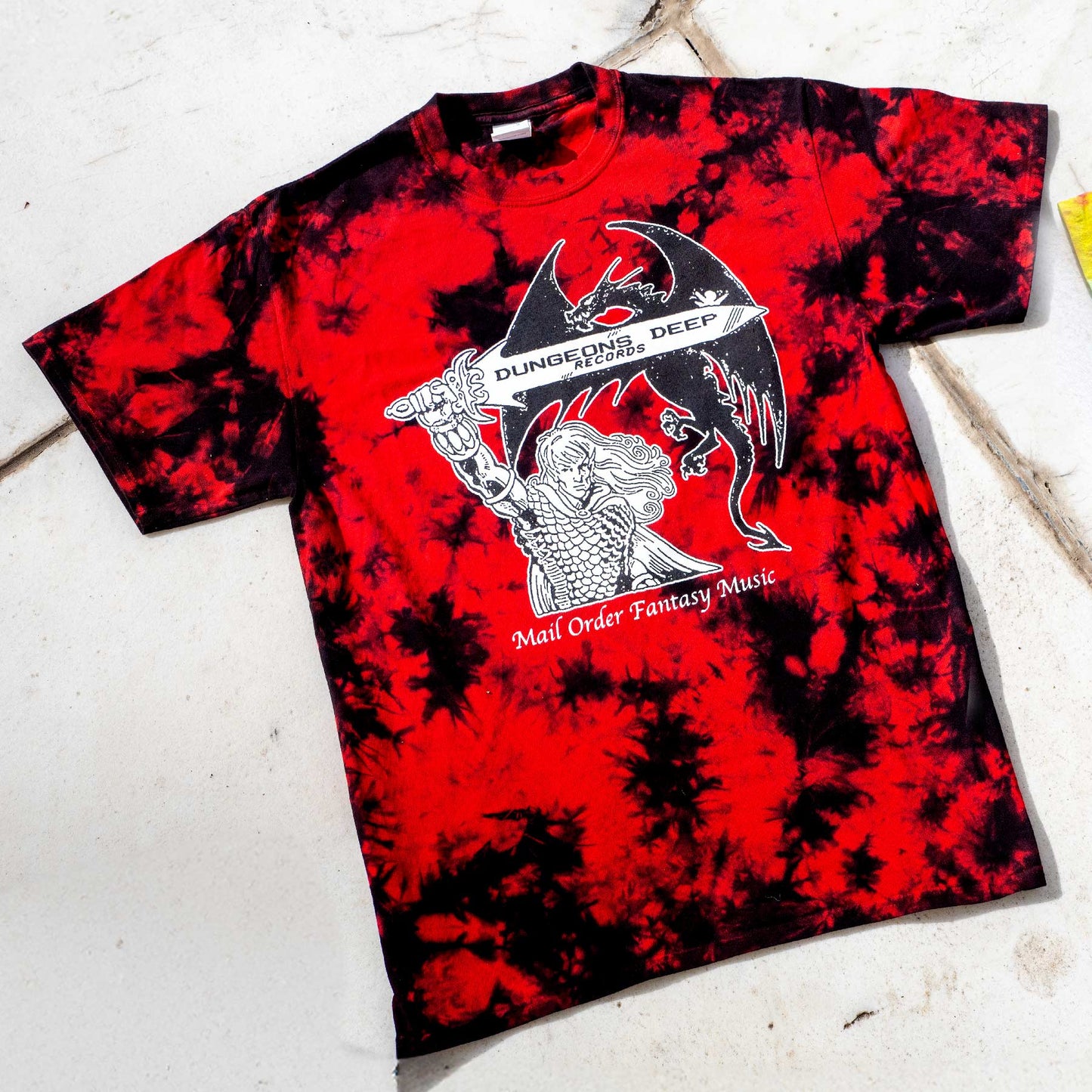 A black and red  tie dye shirt featuring 'Mail Order Fantasy Music' text and illustrations screen printed on the front. The design depicts a man holding a sword with the words "Dungeons Deep Records" inscribed next to a fantasy dragon.