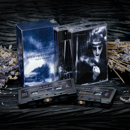 A dark gothic music release including a blue illustrated box, a clear cassette case with a black-and-white portrait insert, and two black cassettes on a dark textured surface with dried lavender props.