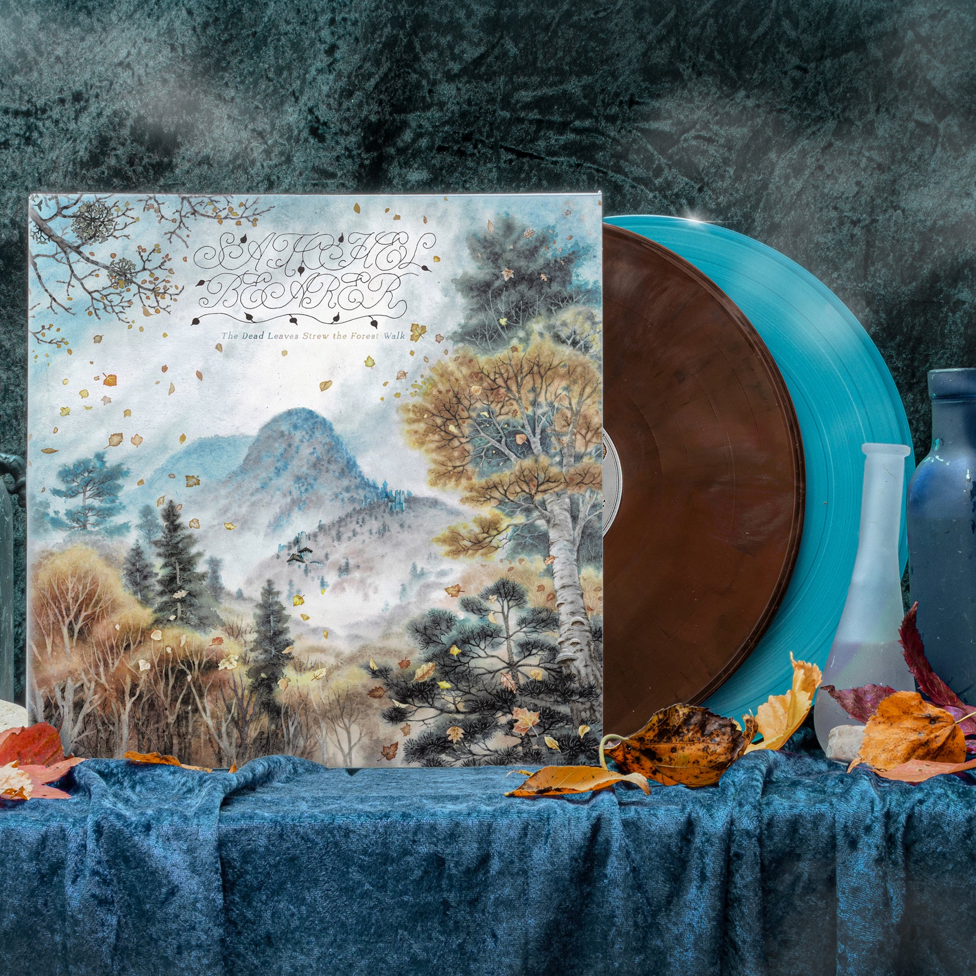 Two vinyl records sitting on a blue velvet backdrop with leaves and glass bottles. One record is brown, the second is light blue.